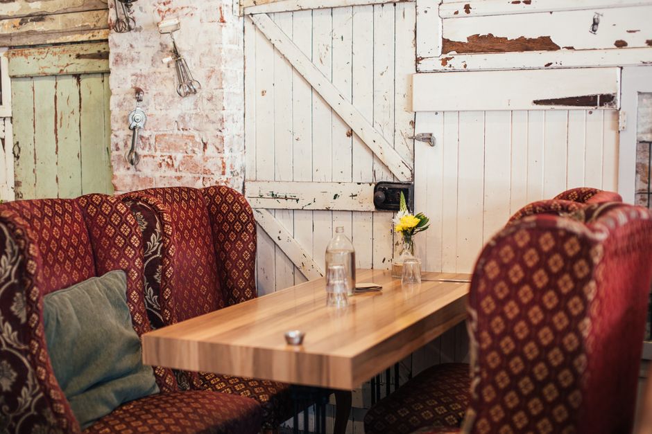 A rustic indoor dining area featuring a polished wooden table set against a weathered white brick wall with peeling paint. The table is adorned with a small glass vase containing yellow flowers and tw